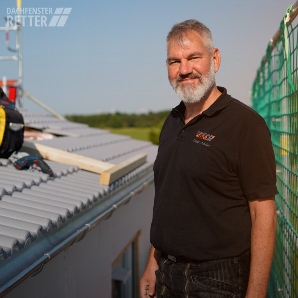 Bild zeigt den Betriebsleiter der DACHFENSTER-RETTER, Oliver Dundiew, im schwarzen Polo-T-Shirt der DACHFENSTER-RETTER, während der Anwesenheit auf einer Baustelle. Es ist ein Fangzaun und ein Dach mit grauen Ziegeln im Hintergrund zu erkennen. 