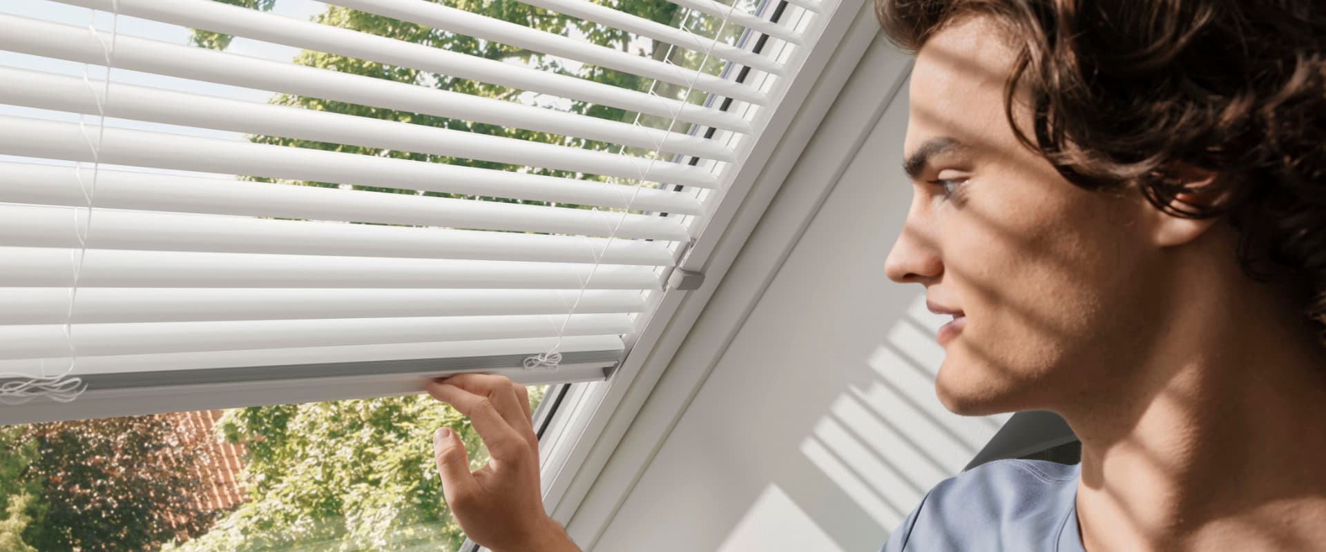 Junger Mann bedient Jalousie an Dachfenster